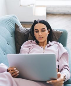 From above young woman in lilac clothes lying on comfortable couch and using netbook in sunlit living room at home